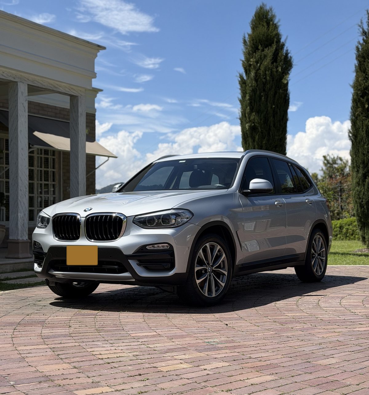 BMW X3 plateado recién lavado frente a una casa, con cielo azul y cipreses